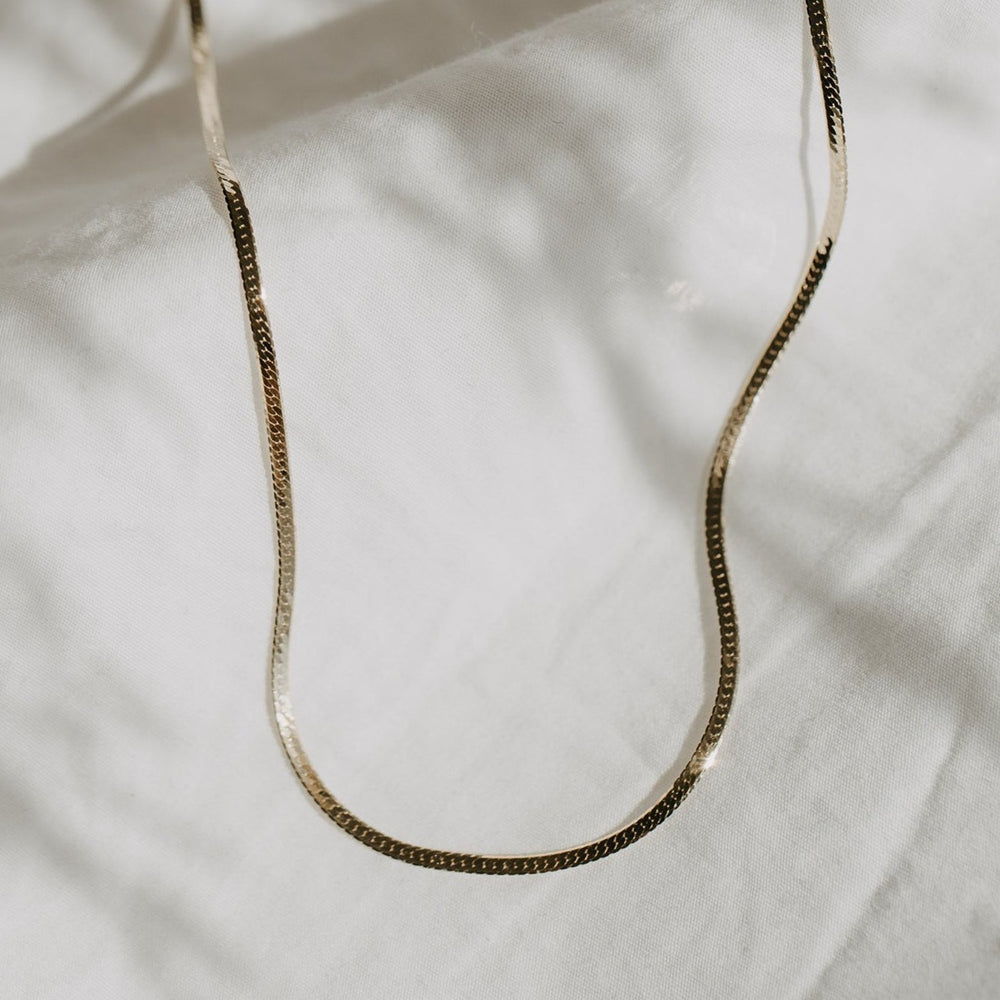 
                      
                        A delicate Thin Herringbone Chain by Pear. rests on a wrinkled, light-colored fabric background. Soft shadows and light play across the surface, highlighting the texture of both the chain and the fabric.
                      
                    