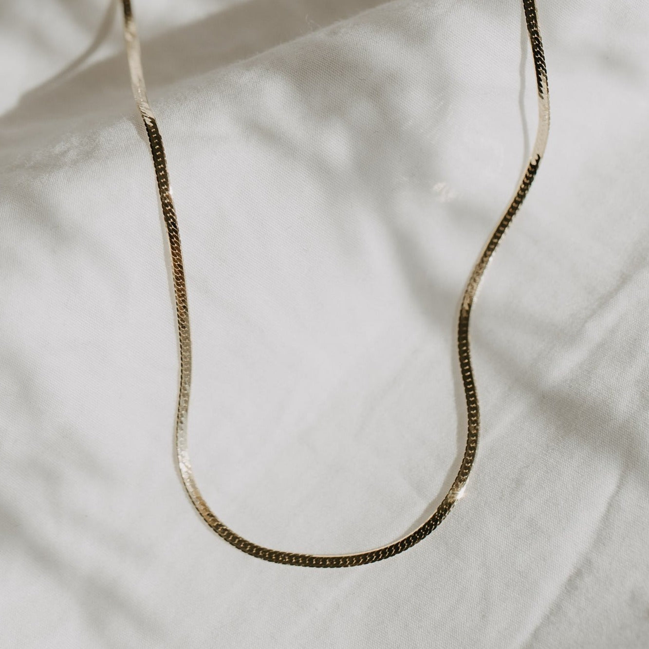 A delicate Thin Herringbone Chain by Pear. rests on a wrinkled, light-colored fabric background. Soft shadows and light play across the surface, highlighting the texture of both the chain and the fabric.