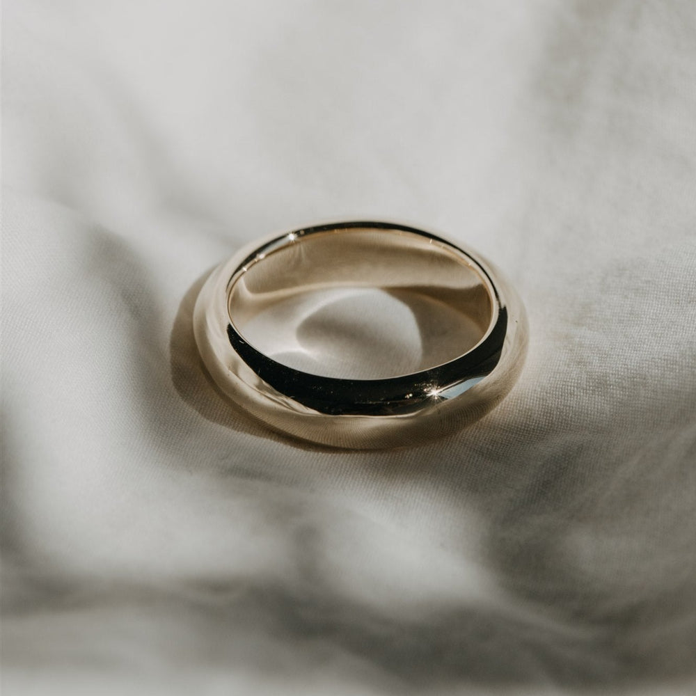 
                      
                        The Pear Heavy Band rests on a soft, white fabric surface. The lighting casts a gentle shadow beneath the solid gold band, and the texture of the fabric is visible, adding depth to the image.
                      
                    