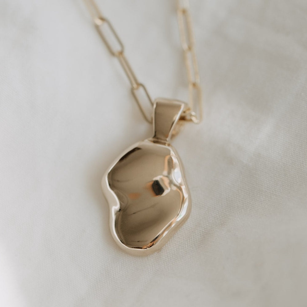 A close-up photo of the Ophelia Necklace by Pear features a solid gold pendant with an abstract, irregular shape hanging from a delicate chain made of elongated links. The pendant's polished surface subtly reflects light, giving it a smooth and shiny appearance. This hand-carved oval treasure is set against a softly blurred background.