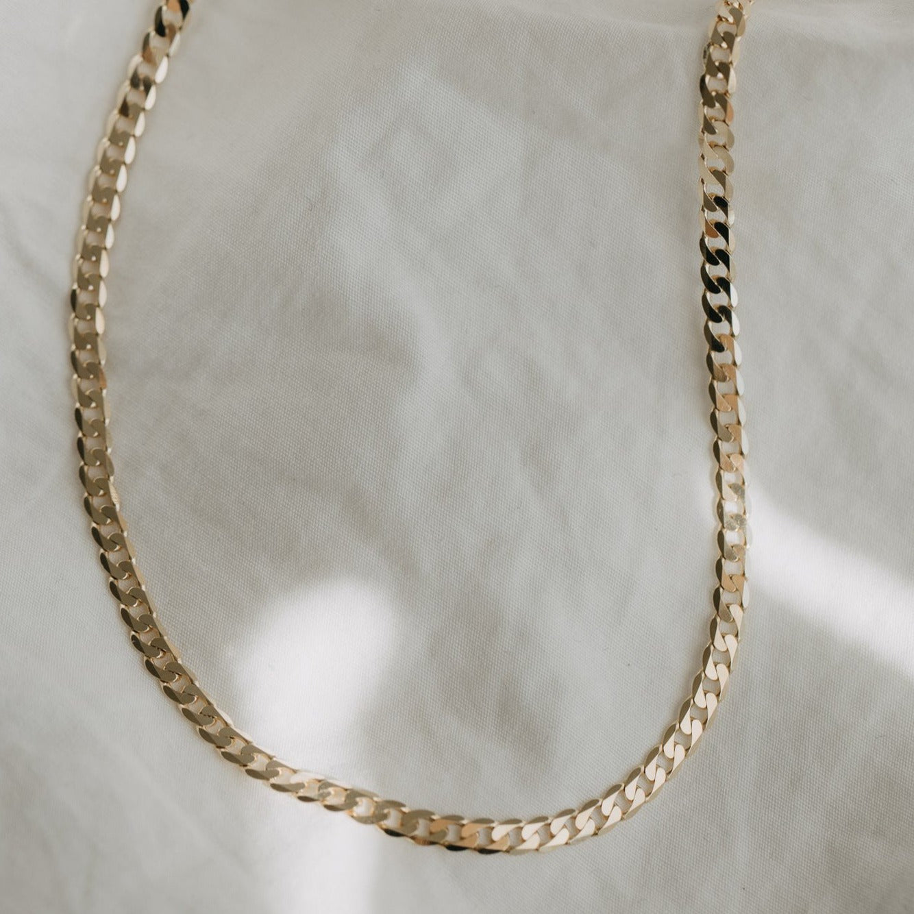 The Pear Curb Chain Necklace laid flat on a soft, textured white cloth background showcases its 14K Solid Yellow Gold construction. The polished and reflective surface of the chain creates subtle light glints, emphasizing its minimalist elegance. Designed for daily wearability, this piece ensures both comfort and style.