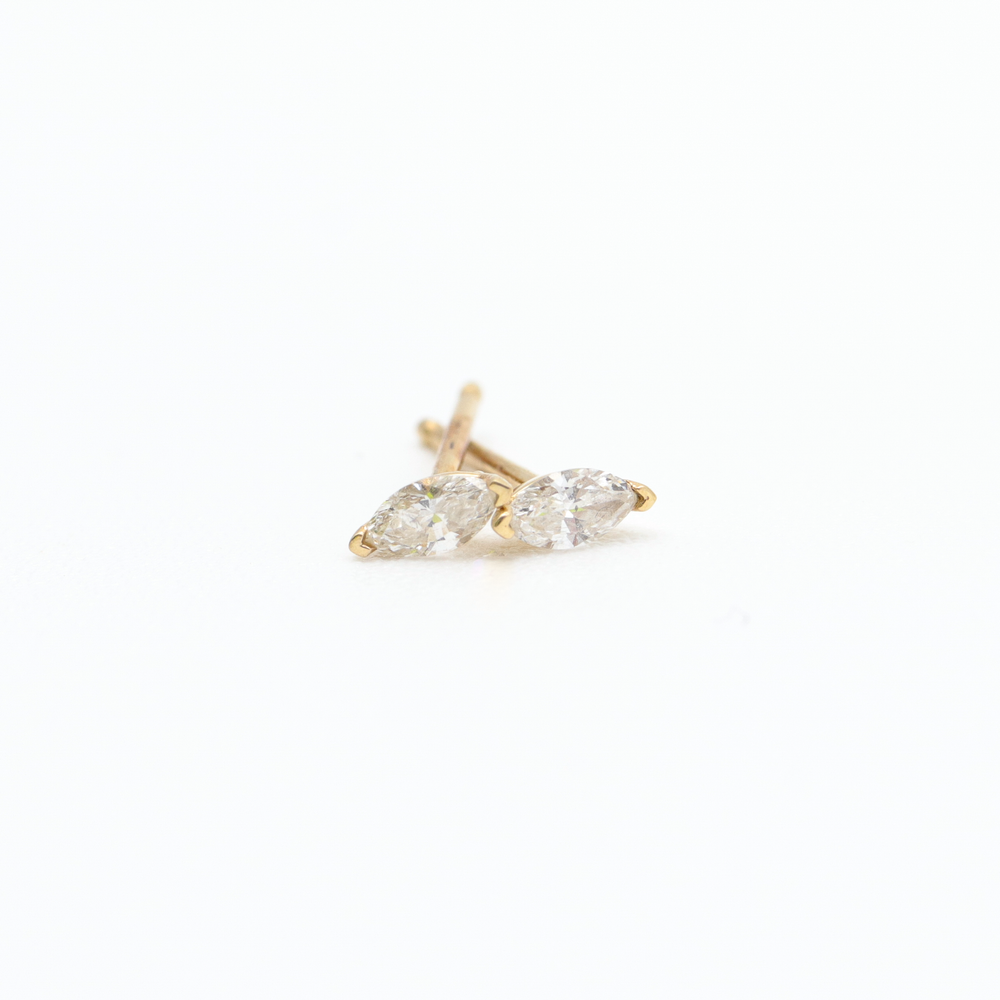 A pair of small, gold Flora Studs by Pear, featuring marquise-cut clear gemstones in a v-prong setting. The earrings are positioned parallel to each other on a white background. Their simple and elegant design is highlighted by the bright lighting.