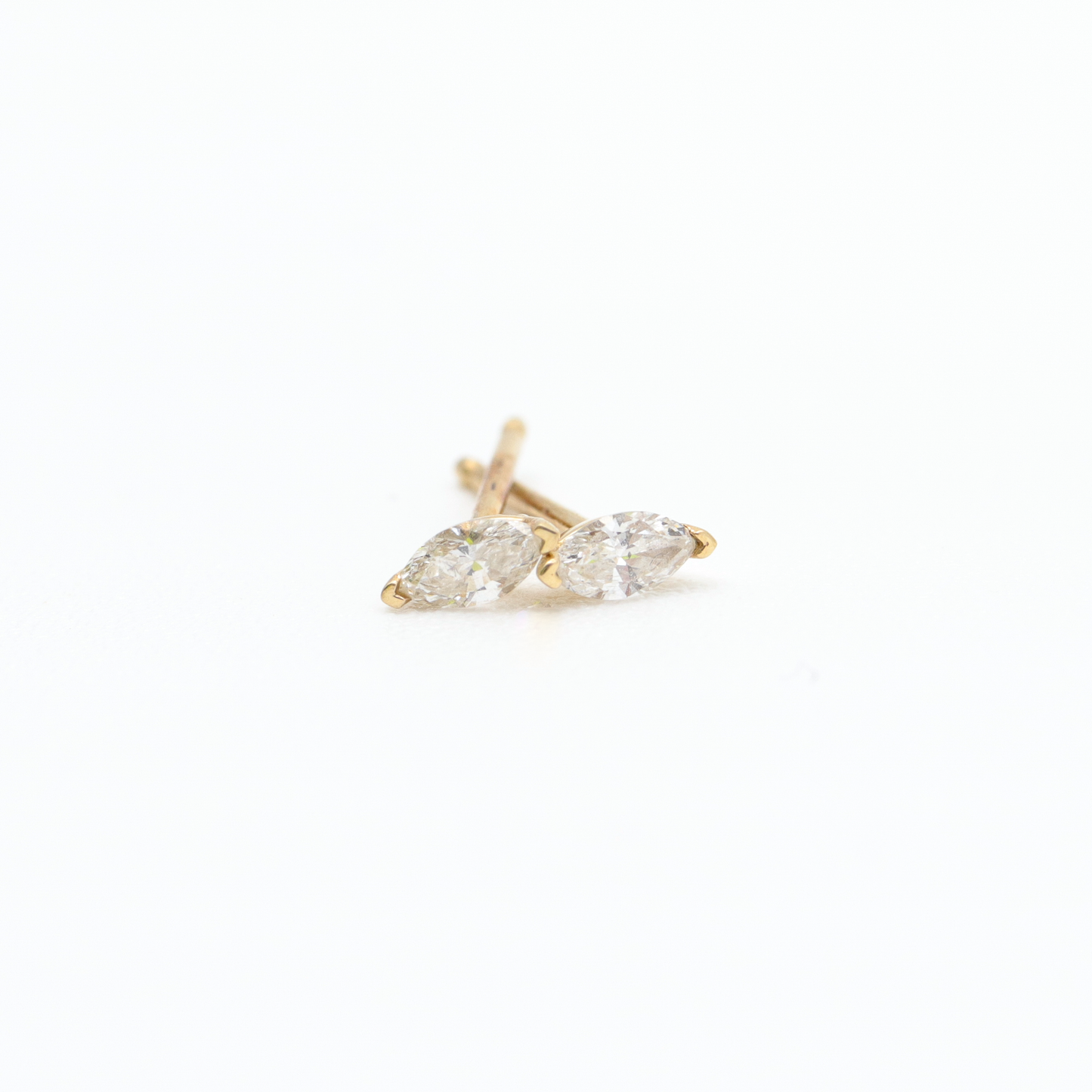 A pair of small, gold Flora Studs by Pear, featuring marquise-cut clear gemstones in a v-prong setting. The earrings are positioned parallel to each other on a white background. Their simple and elegant design is highlighted by the bright lighting.