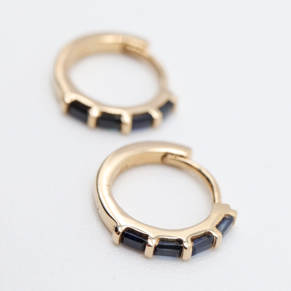 
                      
                        A pair of Sapphire Huggies by Pear., with each gold hoop adorned with five small, dark blue sapphire inlays evenly spaced along the lower half, elegantly displayed on a neutral white background.
                      
                    