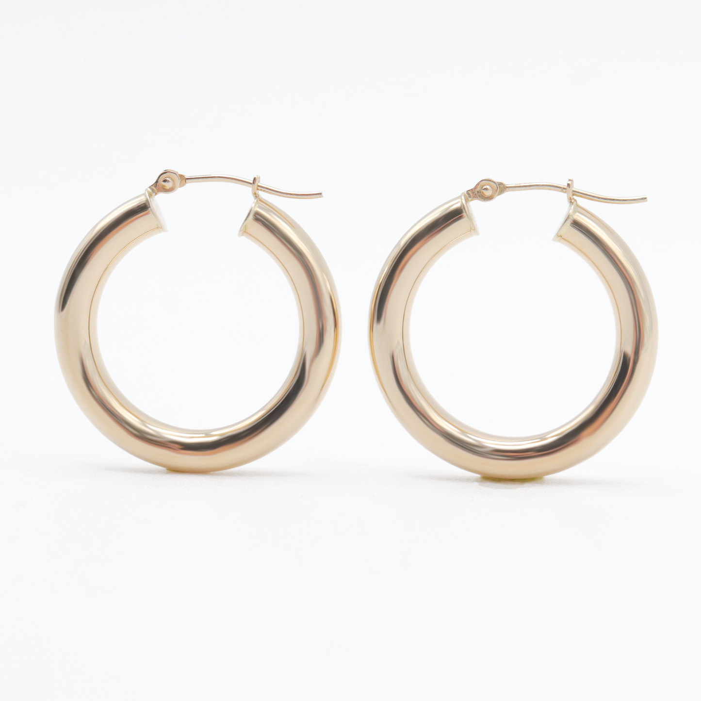 A pair of Pear. Thick Tube Hoops crafted from shiny, solid 14K gold with a polished finish. Featuring a classic circular shape, these earrings are photographed against a plain white background to emphasize their simplicity and elegance. The lightweight latch-back closures are clearly visible at the top, highlighting their practical design.