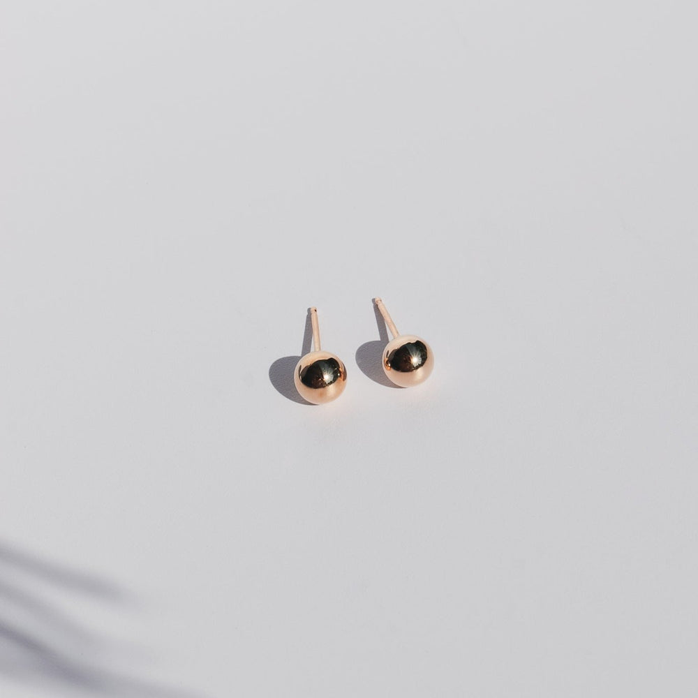 A pair of minimalist The Ball Studs by Pear, featuring 14K gold and round, metallic black stones set in smooth, gold-colored metal. The earrings are elegantly displayed on a clean, light gray surface with a soft shadow beneath them.