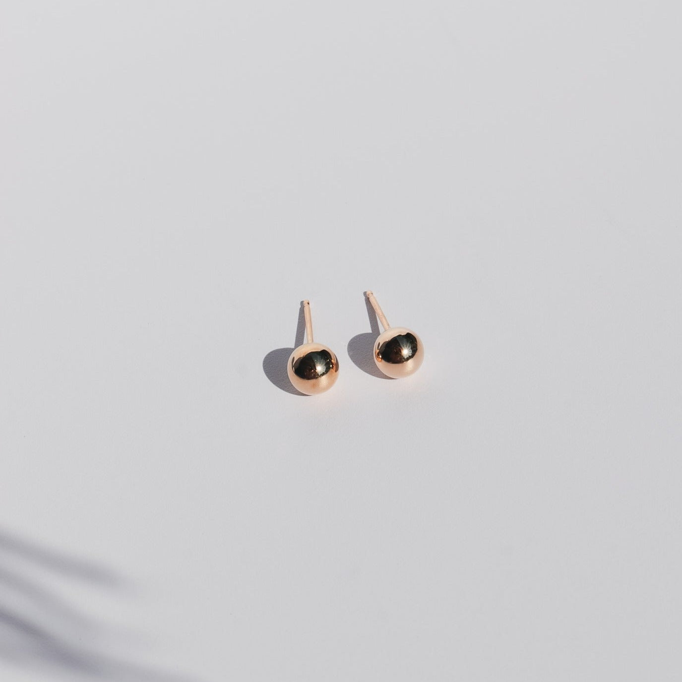 A pair of minimalist The Ball Studs by Pear, featuring 14K gold and round, metallic black stones set in smooth, gold-colored metal. The earrings are elegantly displayed on a clean, light gray surface with a soft shadow beneath them.