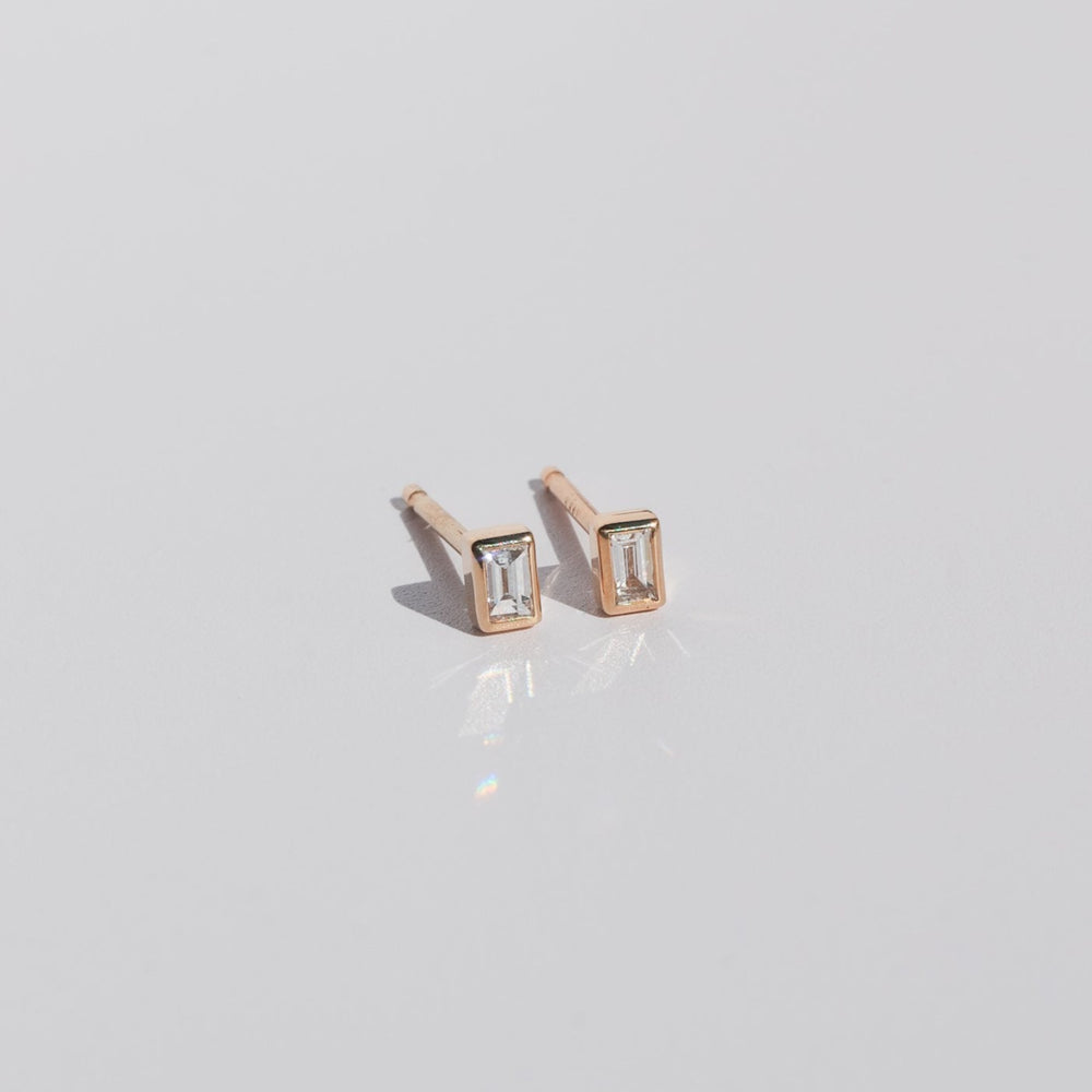 
                      
                        A pair of Diamond Baguette Earrings by Pear with 14K gold settings is displayed on a smooth, reflective surface. The earrings feature a minimalist design and catch the light, casting subtle reflections.
                      
                    