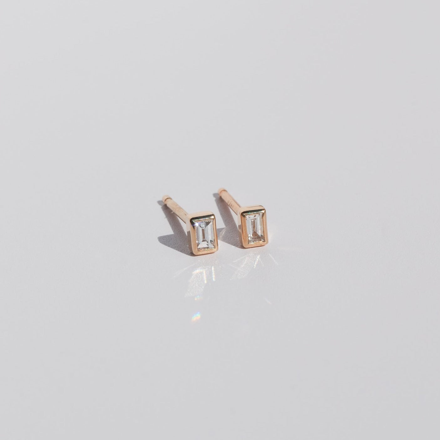 A pair of Diamond Baguette Earrings by Pear with 14K gold settings is displayed on a smooth, reflective surface. The earrings feature a minimalist design and catch the light, casting subtle reflections.