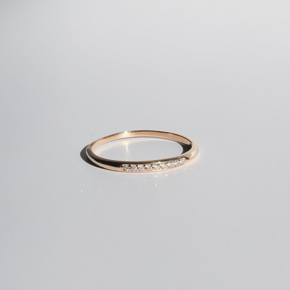 
                      
                        A delicate Esme Diamond Ring by Pear, featuring a simple gold band with a row of small, sparkling diamonds set into the top half, placed on a smooth, light grey surface.
                      
                    