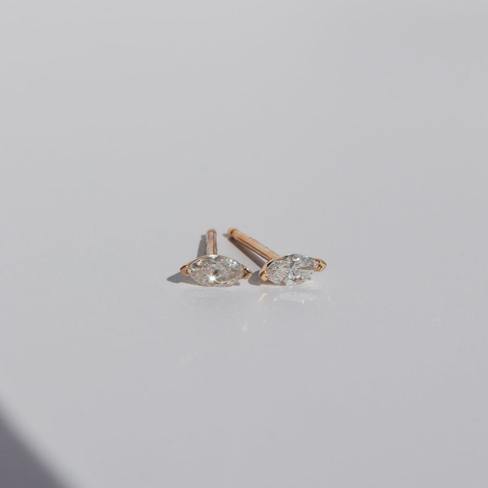 A pair of elegant Flora Studs by Pear featuring oval-shaped clear gemstones in a v-prong setting set in gold. The earrings are placed on a smooth, light-colored surface, showcasing their sparkle and simple yet sophisticated design.